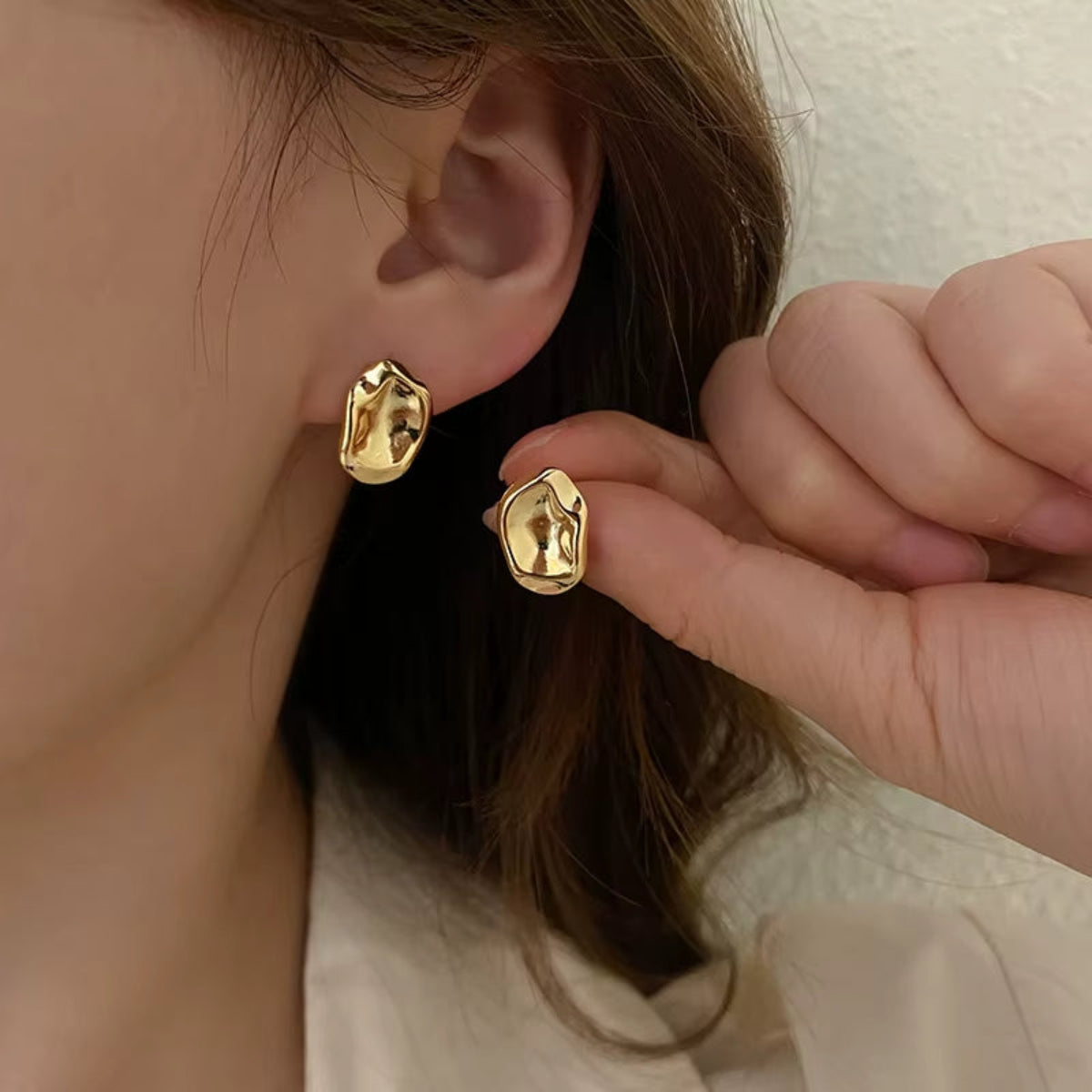 Gold sculptural stud earrings shown on model’s ear for scale and styling